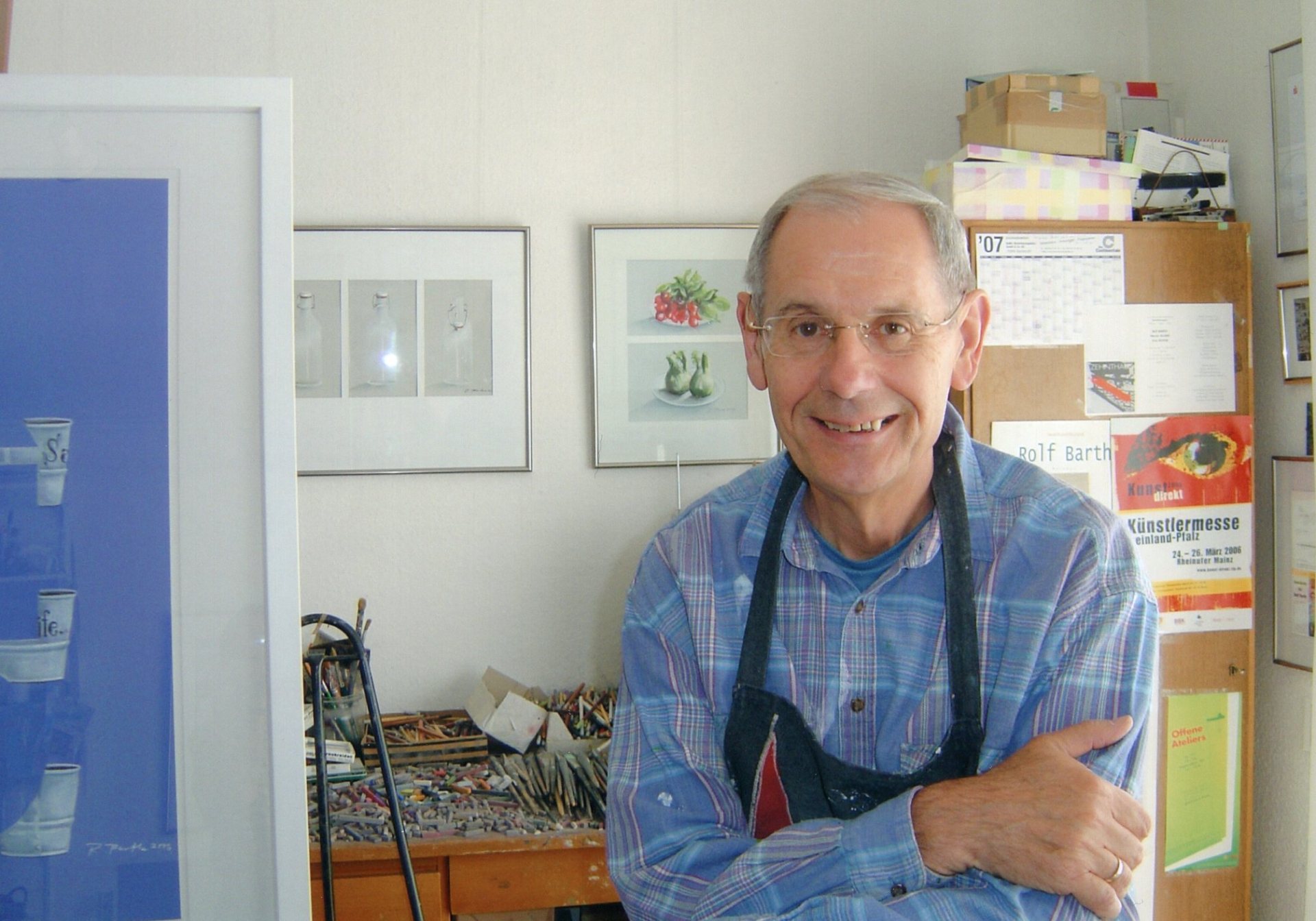 Rolf Barth Blick in ein Atelier; ein Herr in Arbeitskleidung eines Malers blickt lächelnd in die Kamera; imHintergrund sind Bilder und Arbeitsutensilien zu erkennen;