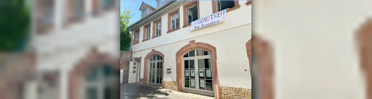 Fassade eines Gebäudes, im Erdgeschoß große Flügeltüren, im Obergeschoß mehrere Fenster, davon sind zwei geöffnet, es hängt ein Banner "Jugendtreff Bad Bergzabern" darunter