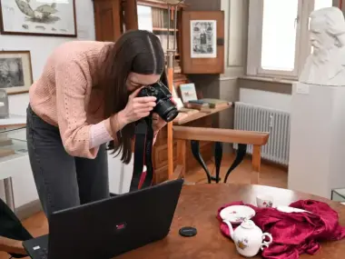 eine Frau in vornübergebeugter Haltung an einem Tisch. Sie hat einen Fotoapparat in der Hand und fotografiert vor ihr stehendes Porzellan. Ein Laptop steht auf dem Tisch.